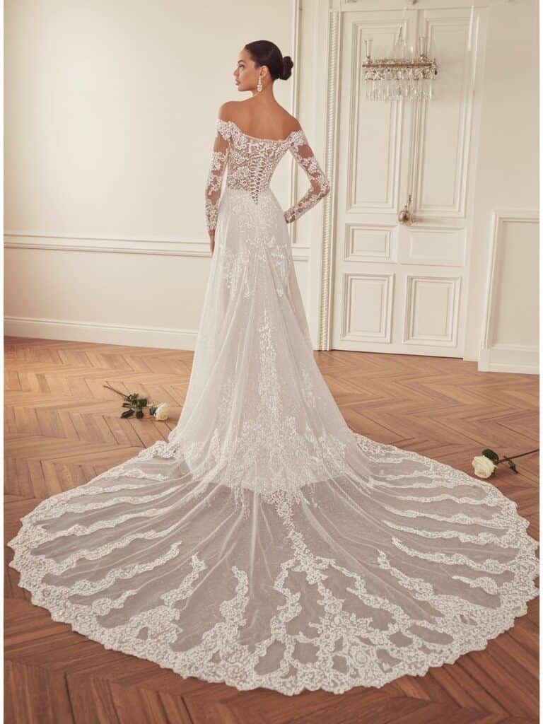 A bride in an off-shoulder, long-sleeved lace wedding dress with a long train, stands back towards the camera in an elegant room with white doors and a parquet floor.