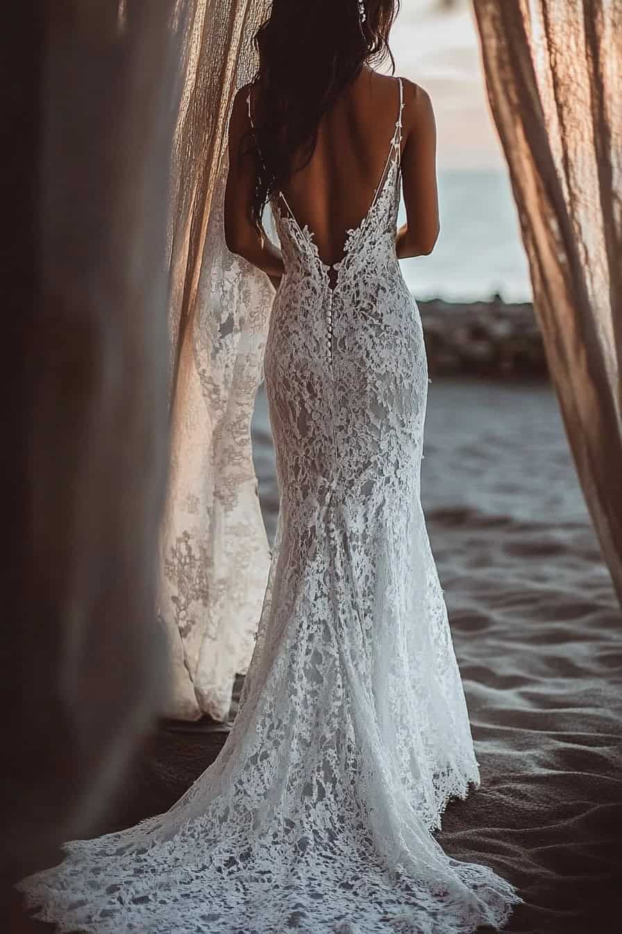 A woman in a lace wedding gown with an open back walks on a sandy beach, framed by draped fabric.