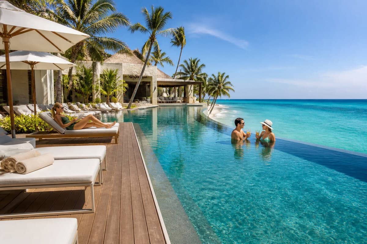 Adults relaxing beside an infinity pool at a luxurious tropical resort overlooking the ocean.
