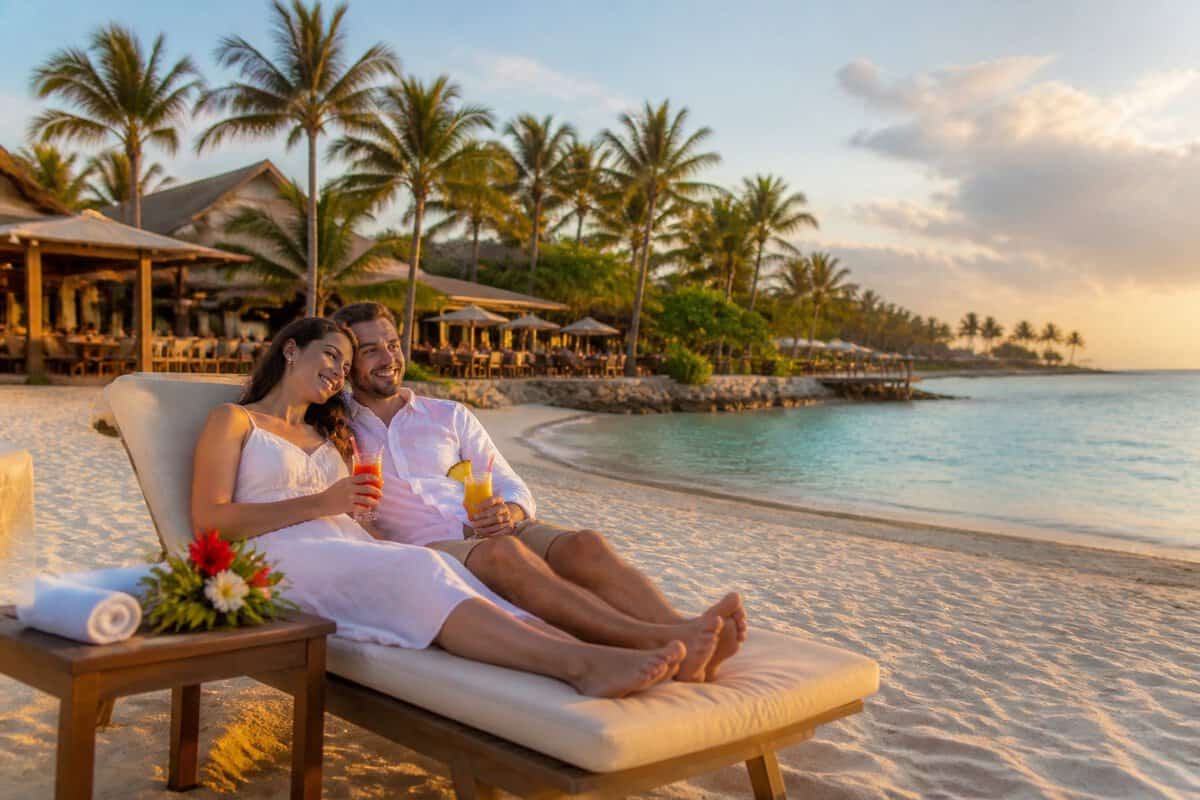 A newlywed couple relaxes together on lounge chairs at a tropical beach resort during sunset.