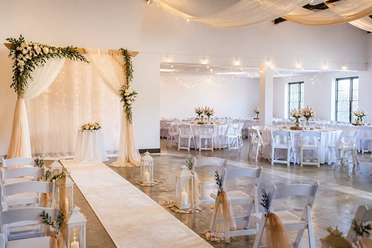 A modest indoor wedding venue decorated with tables, chairs, flowers, and warm lights.