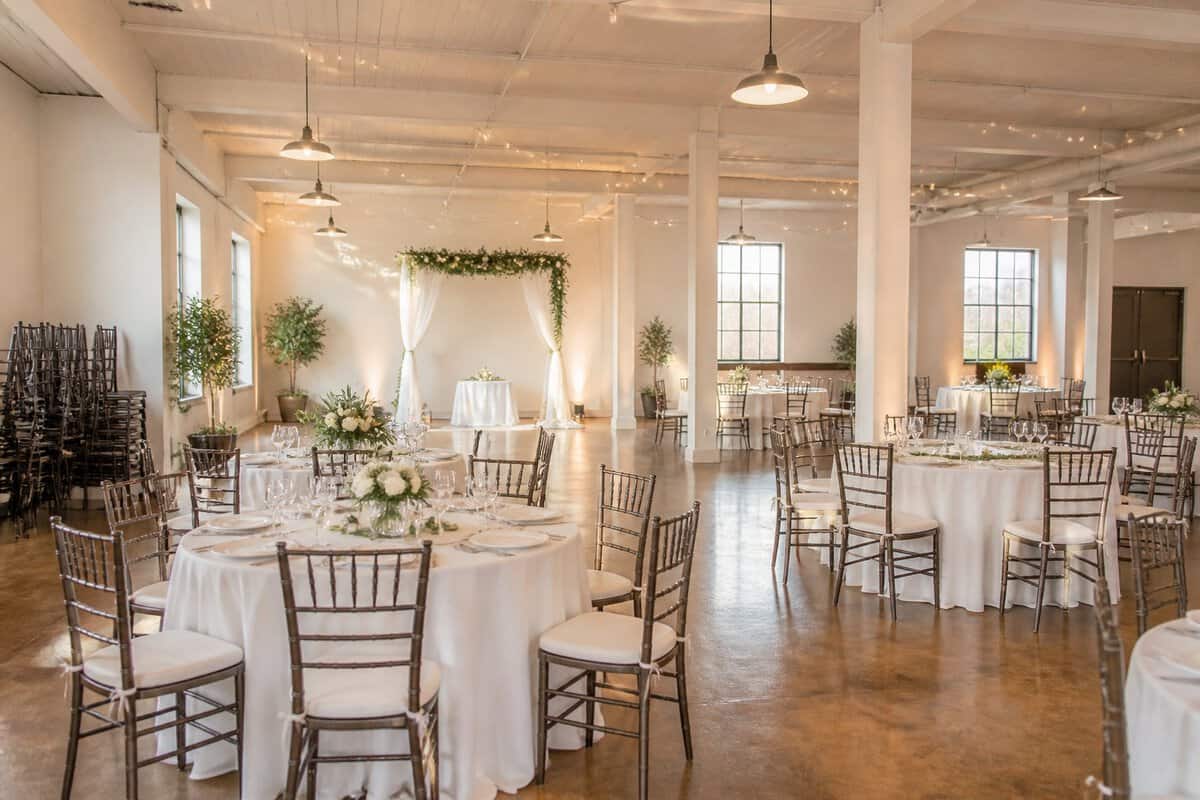 A bright indoor wedding hall with simple elegant tables, greenery, warm lighting, and a small ceremony area.