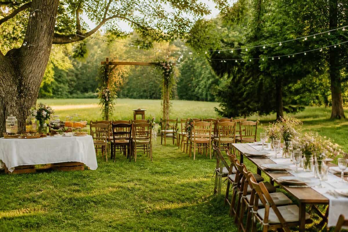 A simple outdoor wedding ceremony and reception setup in a sunny green meadow with wooden chairs, a floral arbor, and decorated tables.