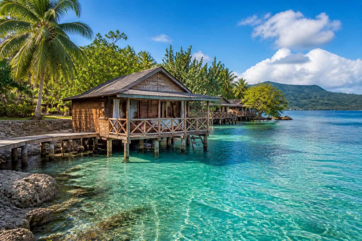 A modest overwater bungalow on stilts above clear turquoise water in a lush Jamaican tropical setting.