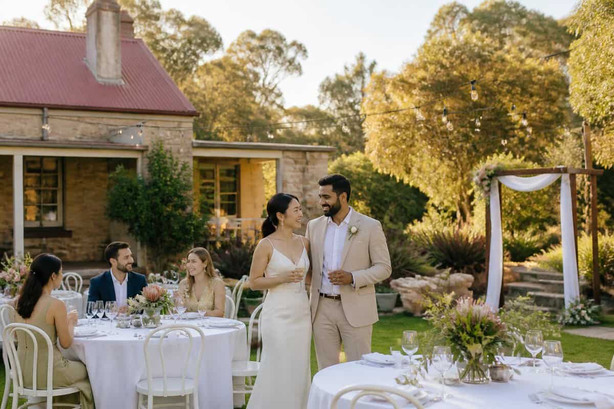 A couple and guests gather at an affordable garden wedding venue with simple tables, flowers, and string lights.