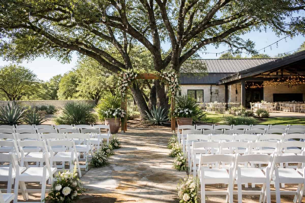 A modest outdoor wedding venue with white chairs, a floral arch, string lights, and a reception area.