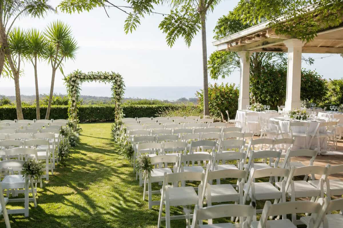 A modestly decorated outdoor wedding venue with white chairs, a floral arch, and a simple reception area in a lush Durban setting.