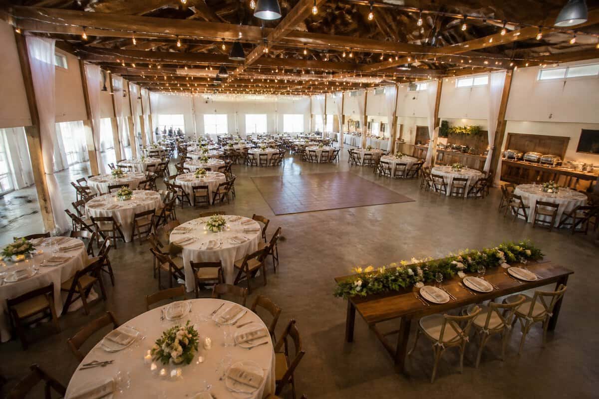 A spacious wedding reception venue arranged with tables, chairs, a dance floor, and simple decorations for a large guest gathering.