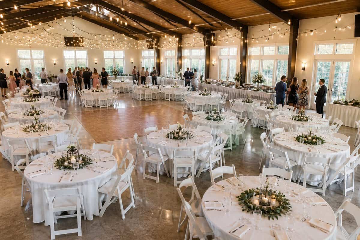 A spacious community hall arranged for a wedding reception with round tables, simple floral centerpieces, warm lights, and guests mingling.