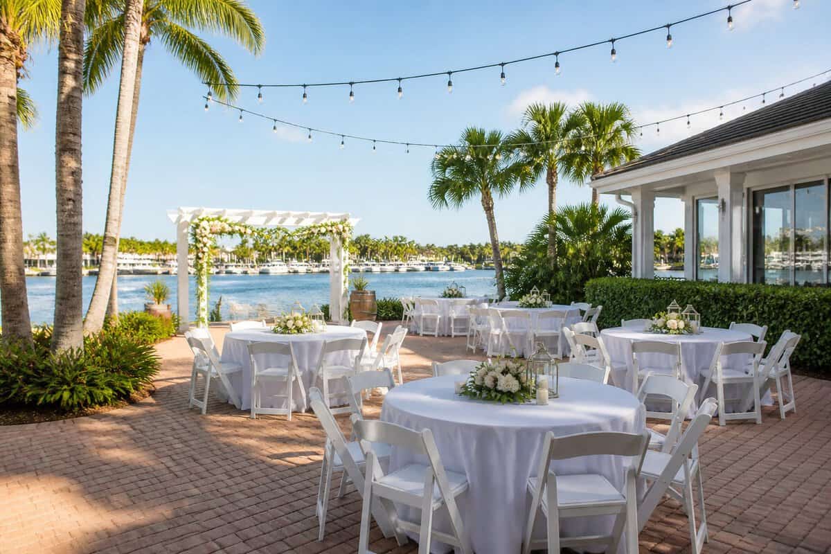 An affordable waterfront wedding venue in Fort Lauderdale with simple floral decorations, arranged tables, palm trees, and calm blue water in the background.