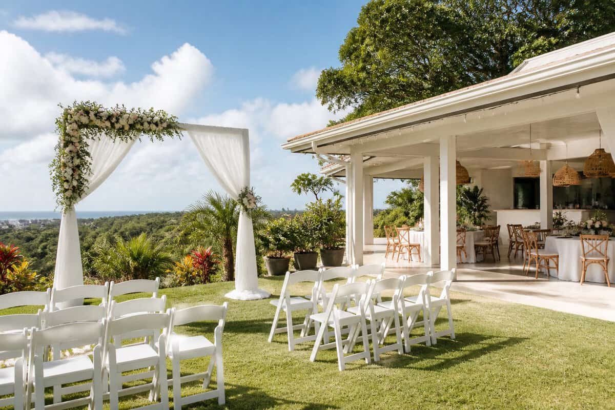 A simple outdoor wedding venue with white chairs, a floral arch, and reception tables surrounded by greenery.