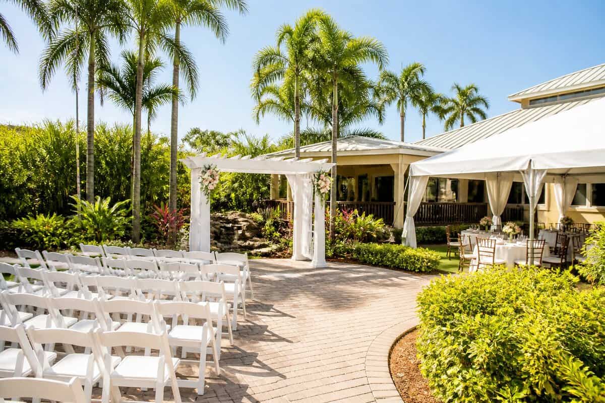 A simple outdoor wedding venue in Fort Myers with a white ceremony arbor, arranged chairs, tropical landscaping, and a covered reception area.