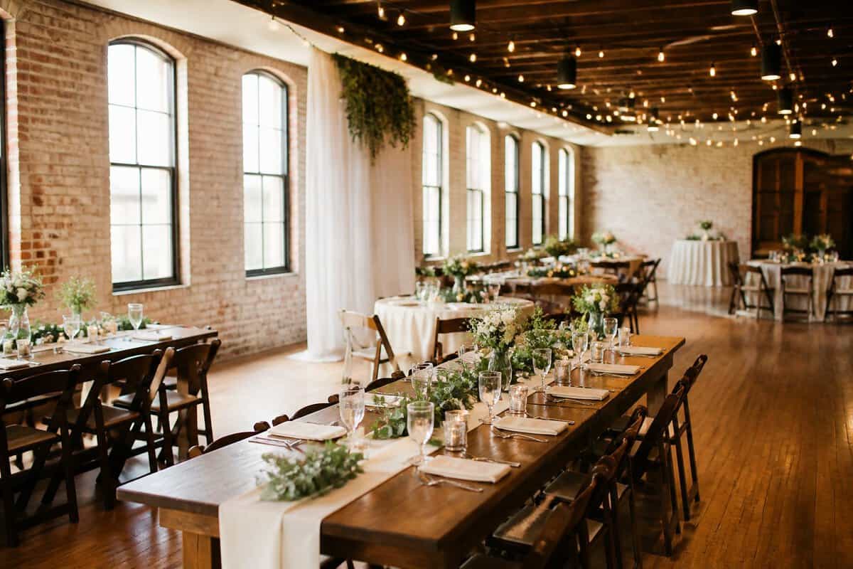 A modest wedding reception room with wooden tables, simple floral centerpieces, and natural daylight.