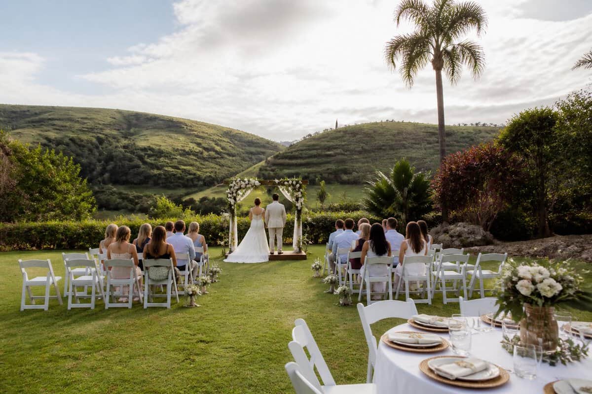 A modest outdoor wedding venue with white chairs, a floral arch, and green hills in the background.