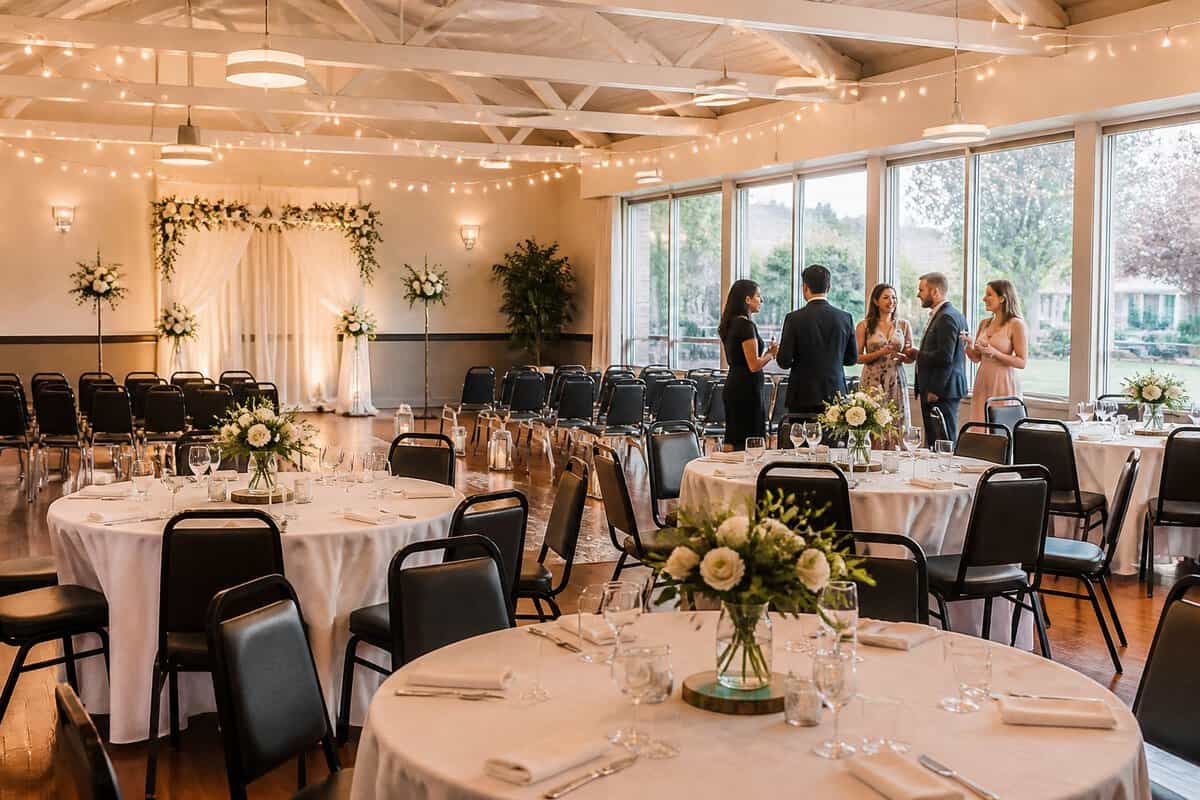 A warmly decorated wedding reception venue with tables, floral centerpieces, string lights, and guests mingling.