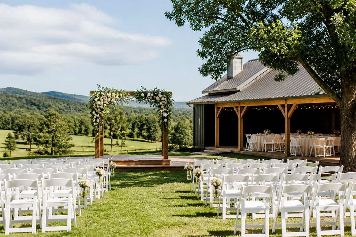 A modest outdoor wedding venue with white chairs, a floral arch, and a reception area surrounded by greenery.