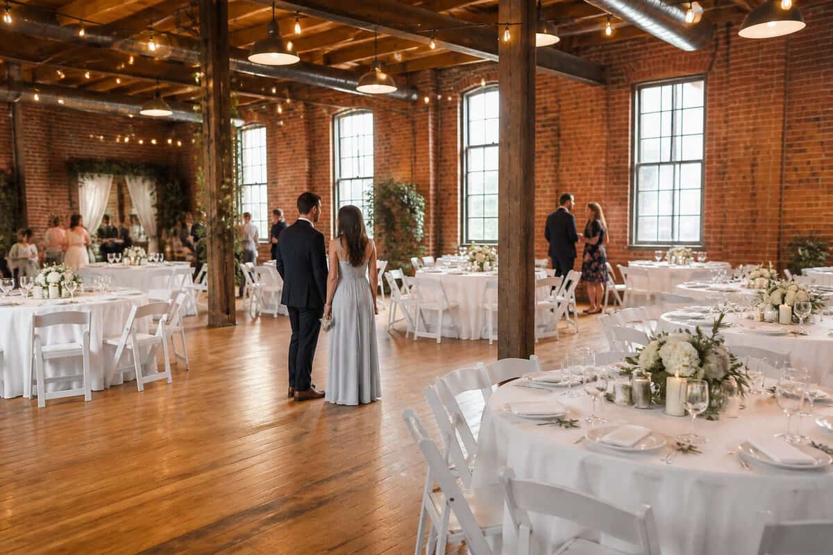 A couple views an affordable wedding reception venue with simple floral tables and warm lighting.
