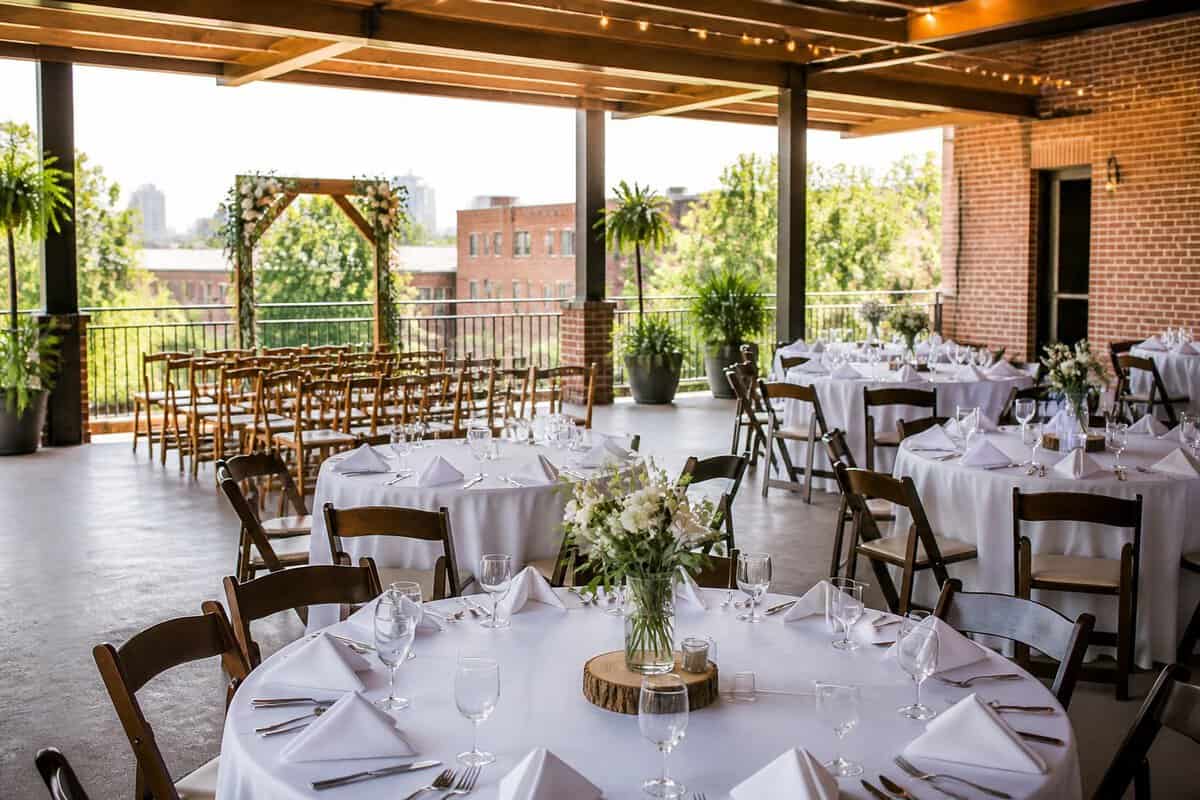 A warmly decorated wedding venue with simple tables, floral centerpieces, string lights, and a small ceremony area.