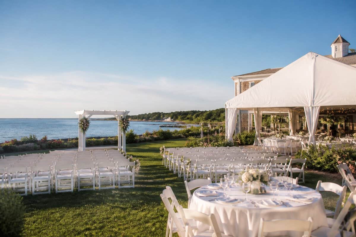 A modest, elegant wedding venue with white chairs, a floral arbor, and a reception tent beside a landscaped waterfront.
