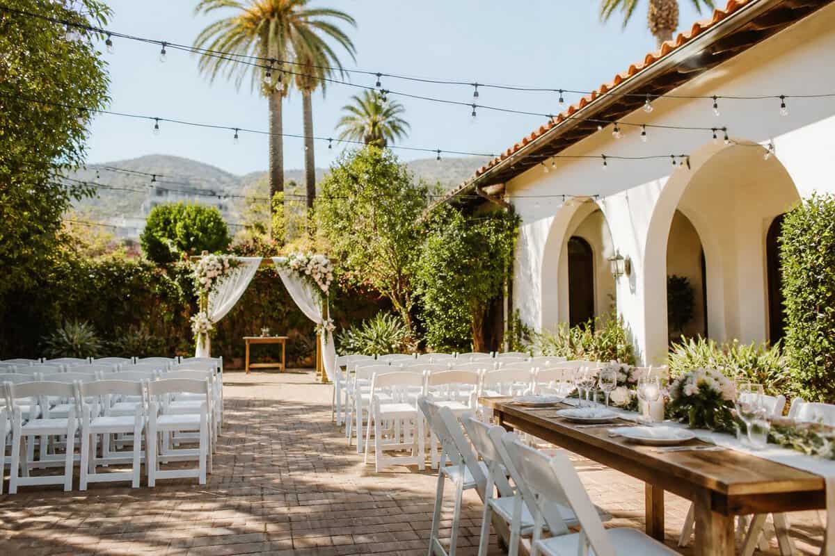 A sunny garden wedding venue with white chairs, floral decorations, string lights, and palm trees.