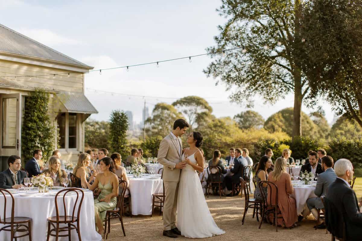 An intimate wedding reception in a leafy Melbourne venue with simple tables, floral decorations, and guests enjoying the outdoor setting.