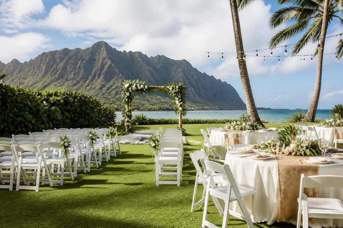 A simple outdoor wedding setup on a grassy lawn near a tropical Oahu beach, with chairs, a floral arch, tables, palm trees, mountains, and ocean.