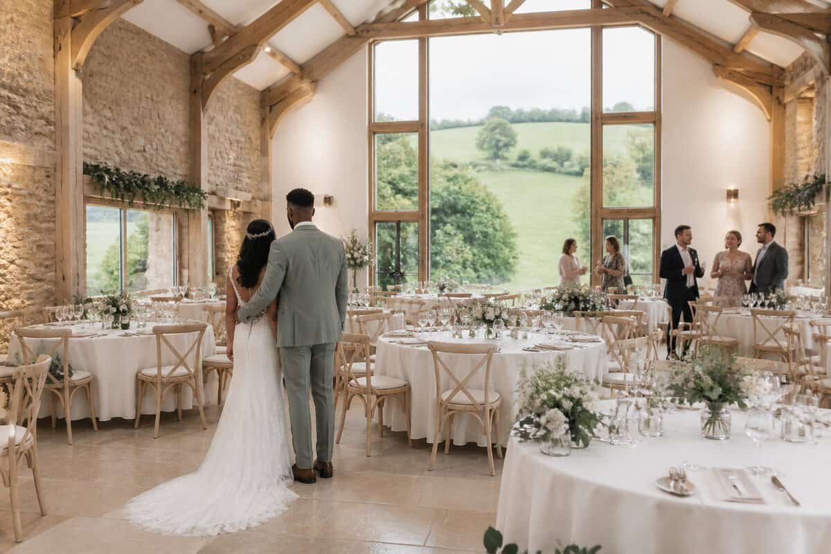 A couple looks over a modestly decorated wedding reception in a bright rustic venue.