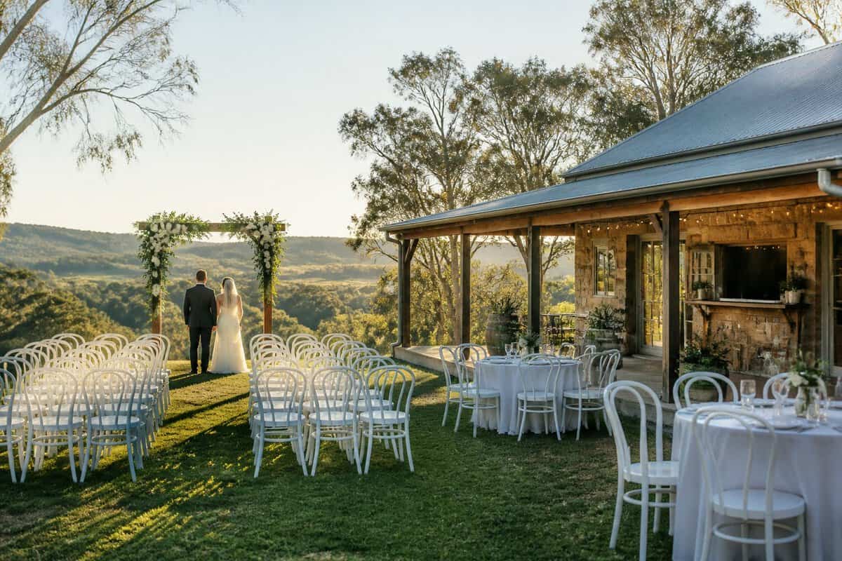 A simple outdoor wedding venue with white chairs, a floral arch, and reception tables in a green countryside setting.