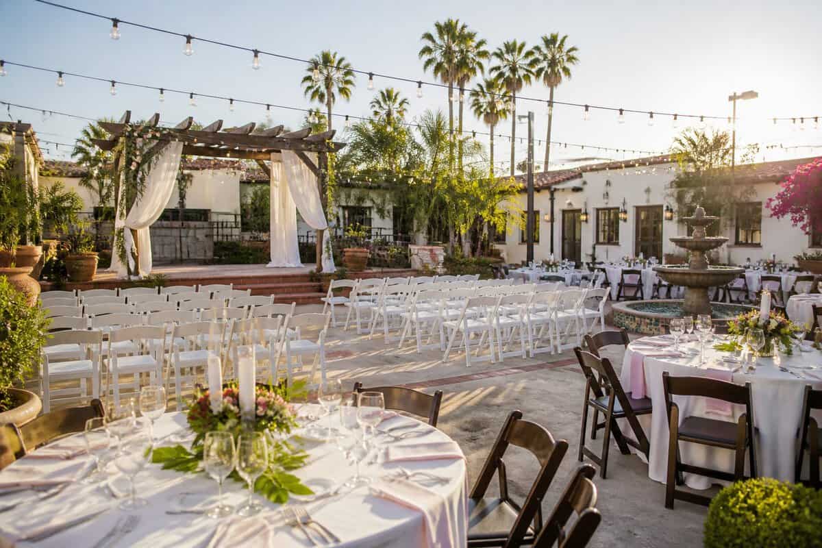 A sunlit garden wedding venue with white chairs, a floral arbor, string lights, and palm trees.