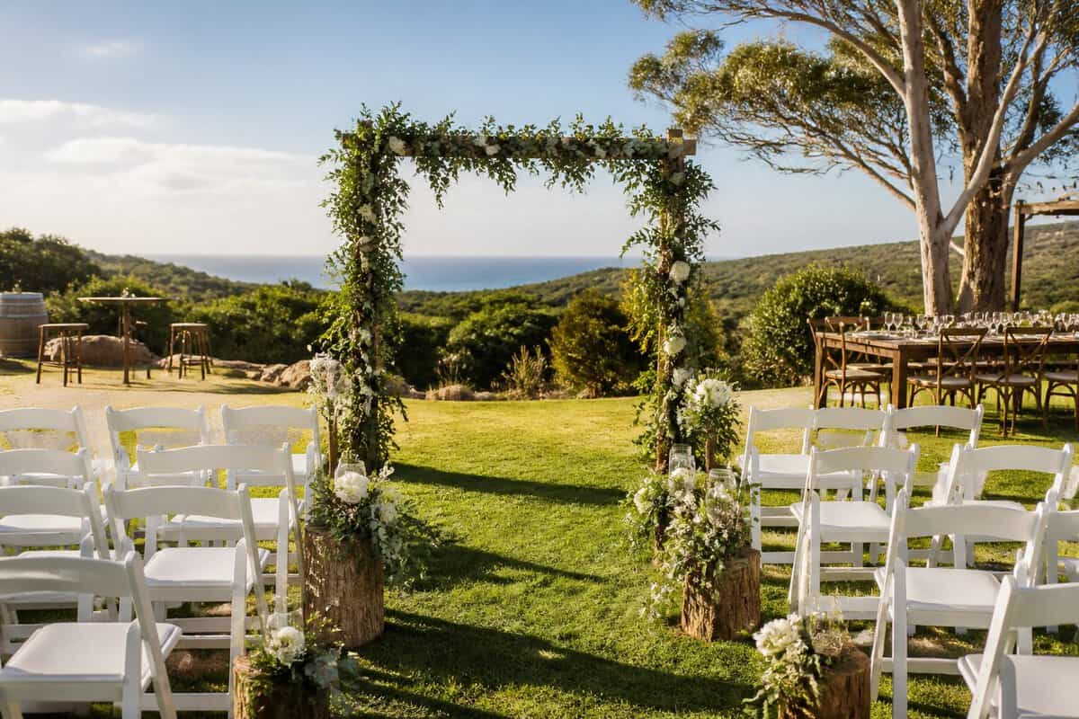 An intimate outdoor wedding ceremony setup with white chairs, flowers, and rustic tables in a sunny garden.