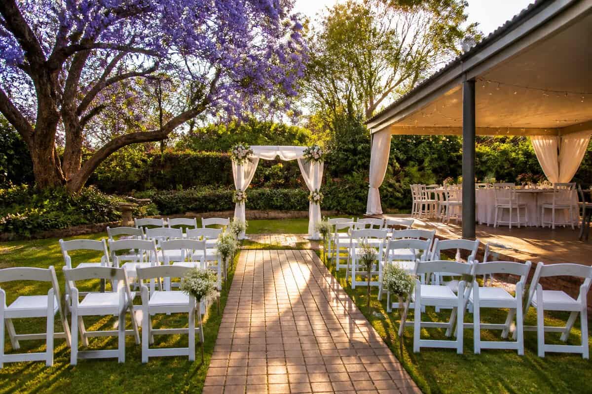 A modest outdoor wedding venue in a lush Pretoria garden with white chairs, simple floral decor, and a covered reception area.