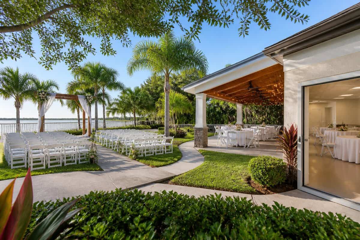 An outdoor wedding ceremony setup with white chairs, simple floral decorations, and a nearby reception area at a landscaped Florida venue.