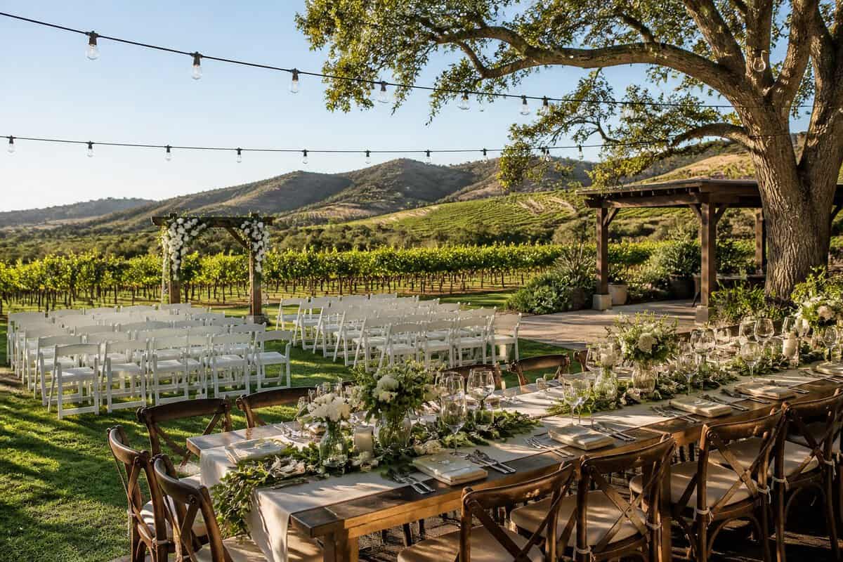 An elegant outdoor wedding venue with white chairs, a floral arch, reception tables, and vineyard hills in the background.