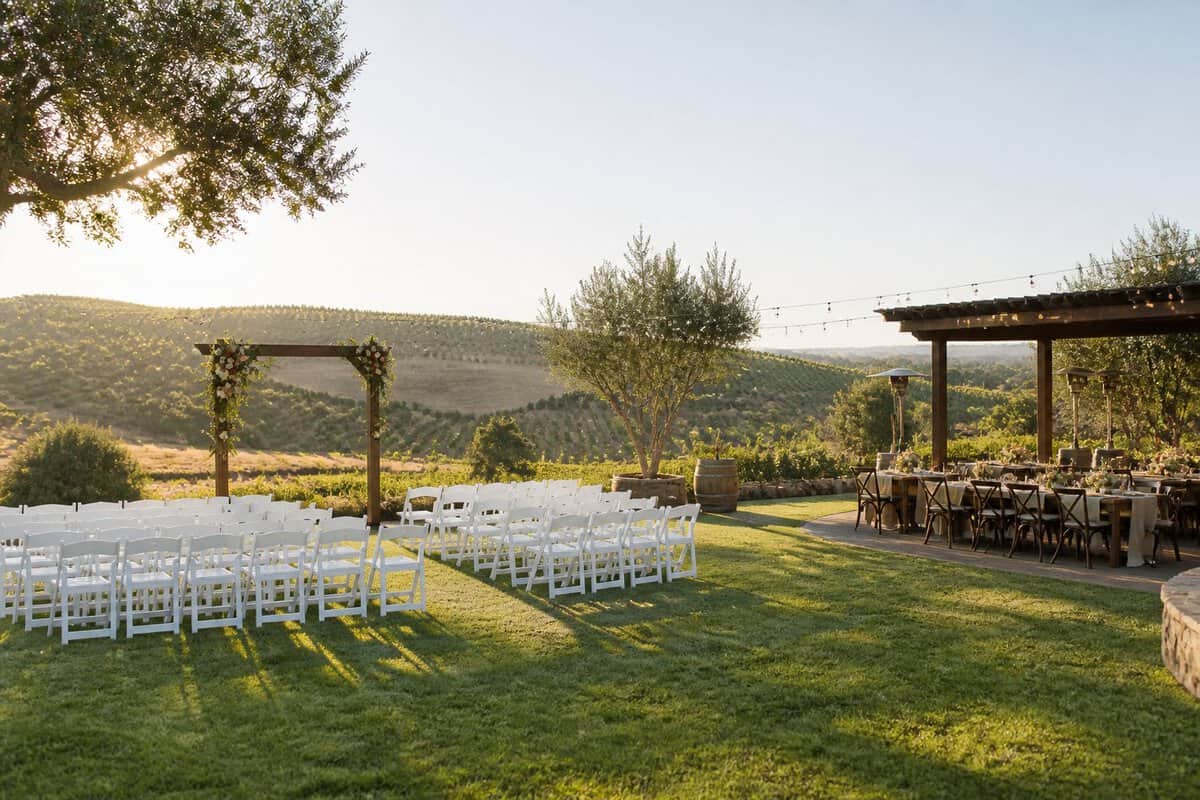 A modest outdoor wedding venue with white chairs, a decorated arbor, and vineyard hills in the background.