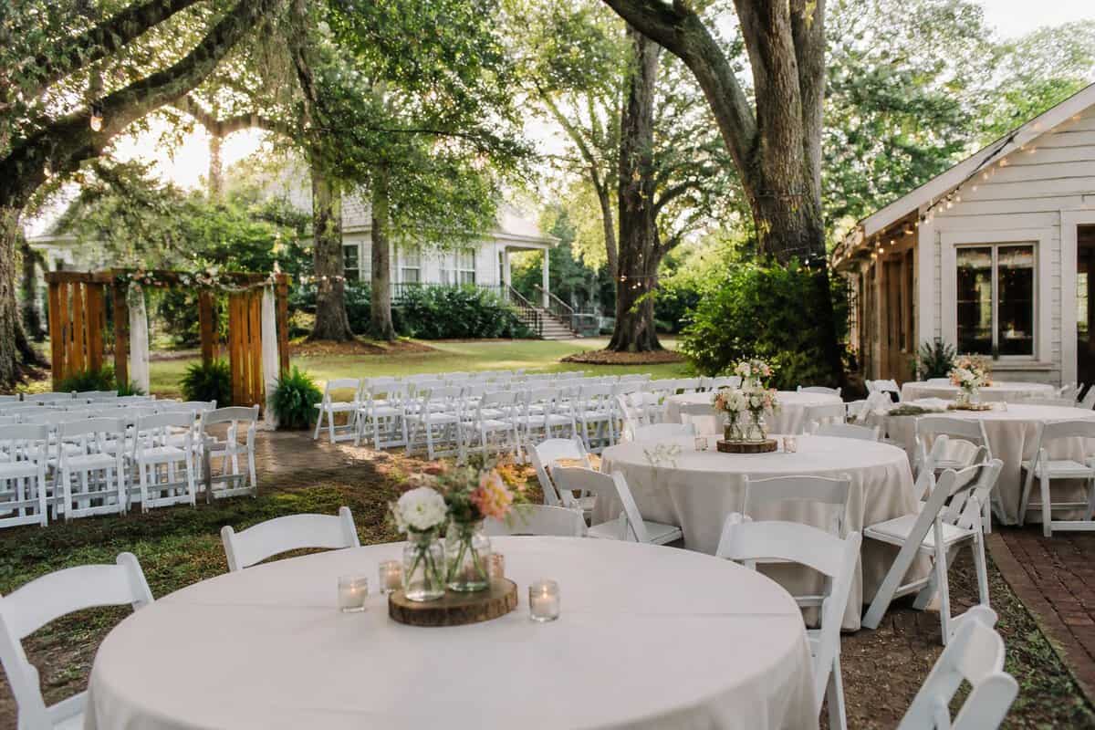 A simple outdoor wedding venue with white chairs, a decorated ceremony arch, and tables beneath oak trees.