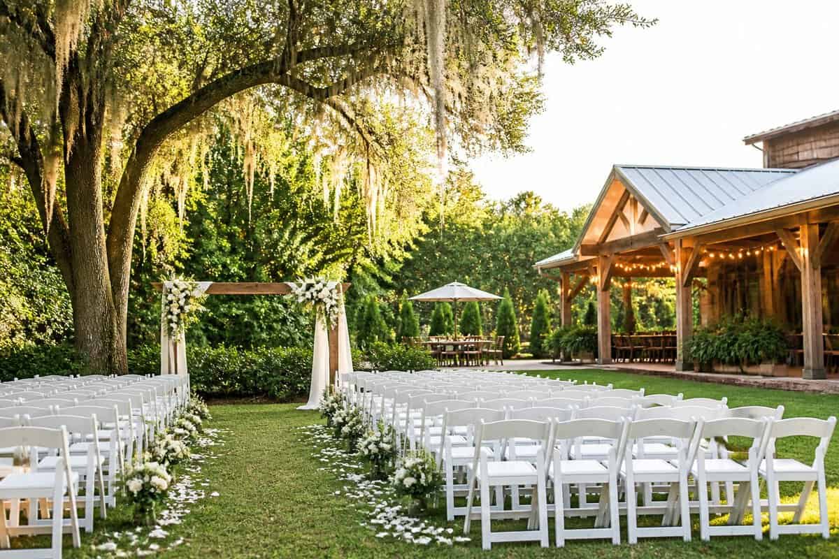 A charming outdoor wedding venue with simple white chairs, a floral aisle, and a wooden ceremony arch surrounded by greenery.