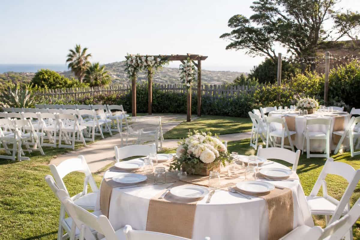 A simple outdoor wedding ceremony and reception setup at a landscaped coastal venue with chairs, tables, flowers, and hills in the background.