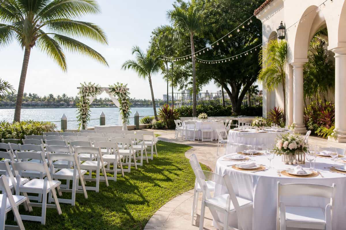 An outdoor wedding venue with white chairs, a floral arch, palm trees, and simple reception tables.