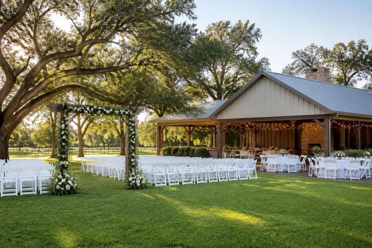 A charming outdoor wedding venue with white chairs, a flower-decorated arbor, and a reception pavilion beneath oak trees.