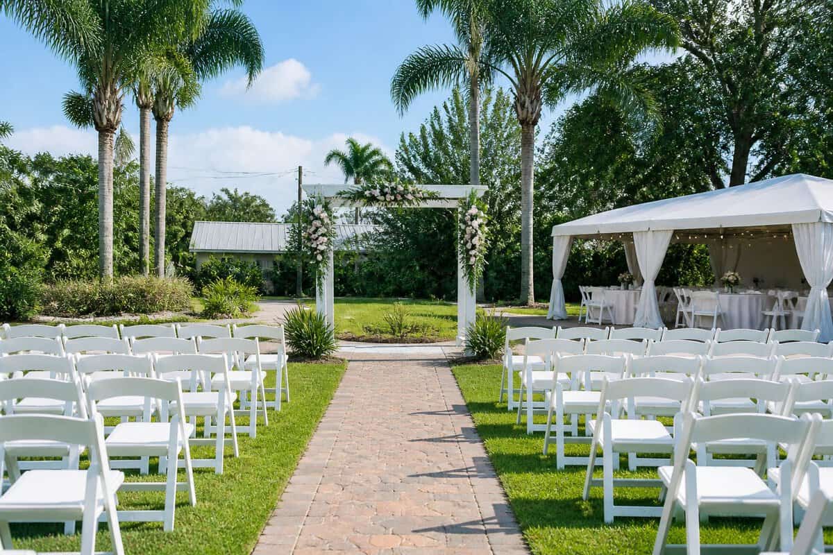 A simple outdoor wedding venue with a flower-decorated arch, white chairs, and a covered reception area surrounded by greenery.