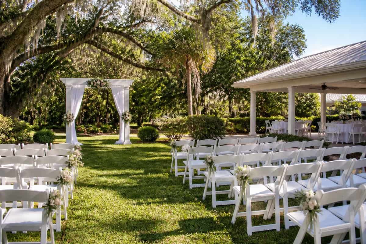 A simple outdoor wedding venue with white chairs, a floral arbor, and a covered reception area surrounded by greenery.