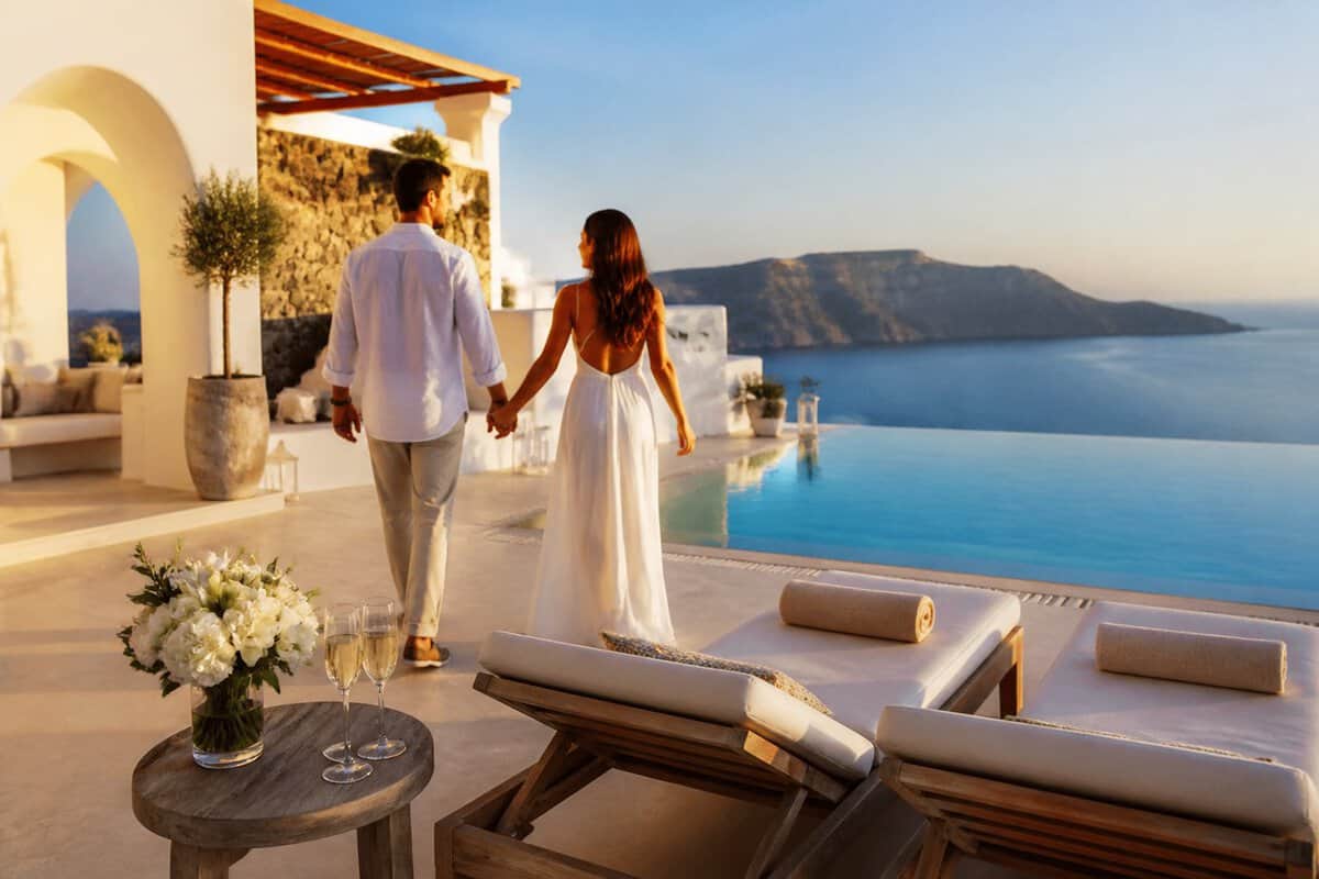 Newlywed couple walking beside a luxurious Greek seaside resort overlooking the blue Aegean Sea.