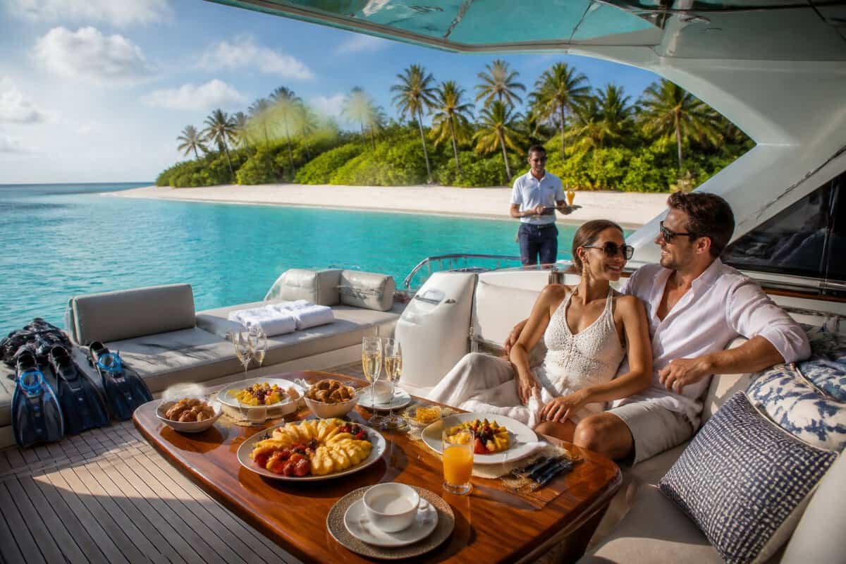 A newlywed couple relaxes on a luxury yacht surrounded by tropical water, breakfast, drinks, and snorkeling equipment.