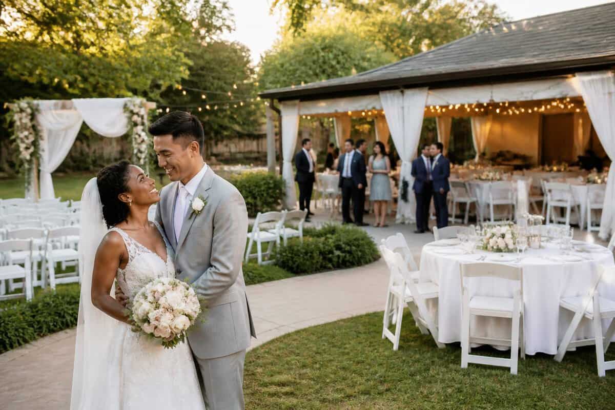 A newlywed couple stands at an outdoor wedding venue with decorated seating, floral arrangements, and guests nearby.