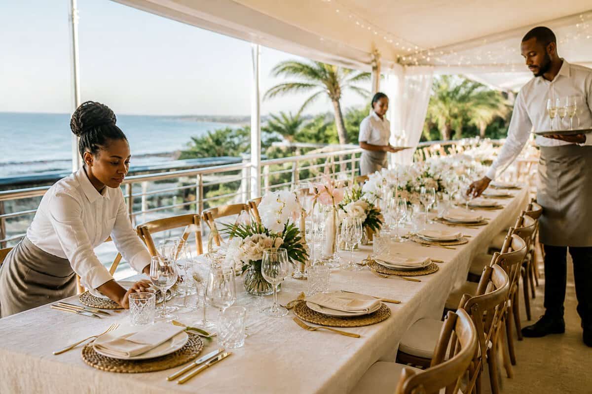 An elegant outdoor wedding reception with a decorated dining table, venue staff, and a coastal garden setting.
