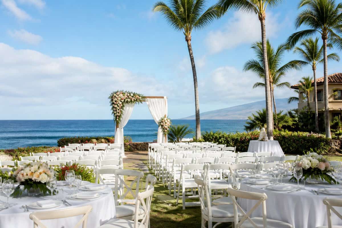 An elegant tropical wedding ceremony setup at a Maui oceanfront resort with white chairs, flowers, palm trees, and reception tables.