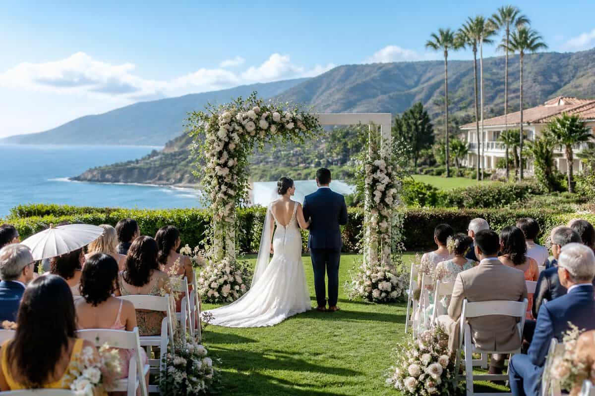 A newlywed couple stands near an outdoor wedding ceremony overlooking a scenic resort landscape, with diverse guests in the background.