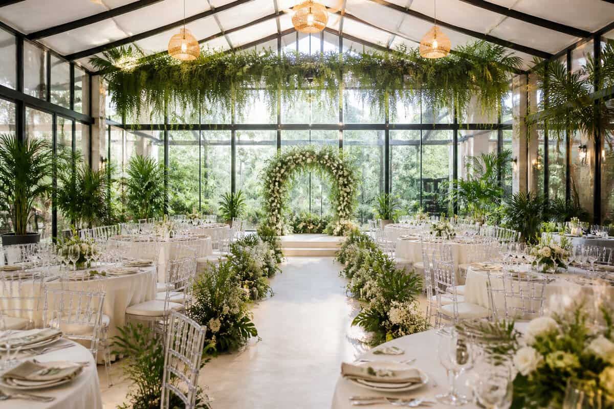 An elegant indoor garden hall arranged for a wedding reception, filled with greenery, flowers, tables, and chairs.