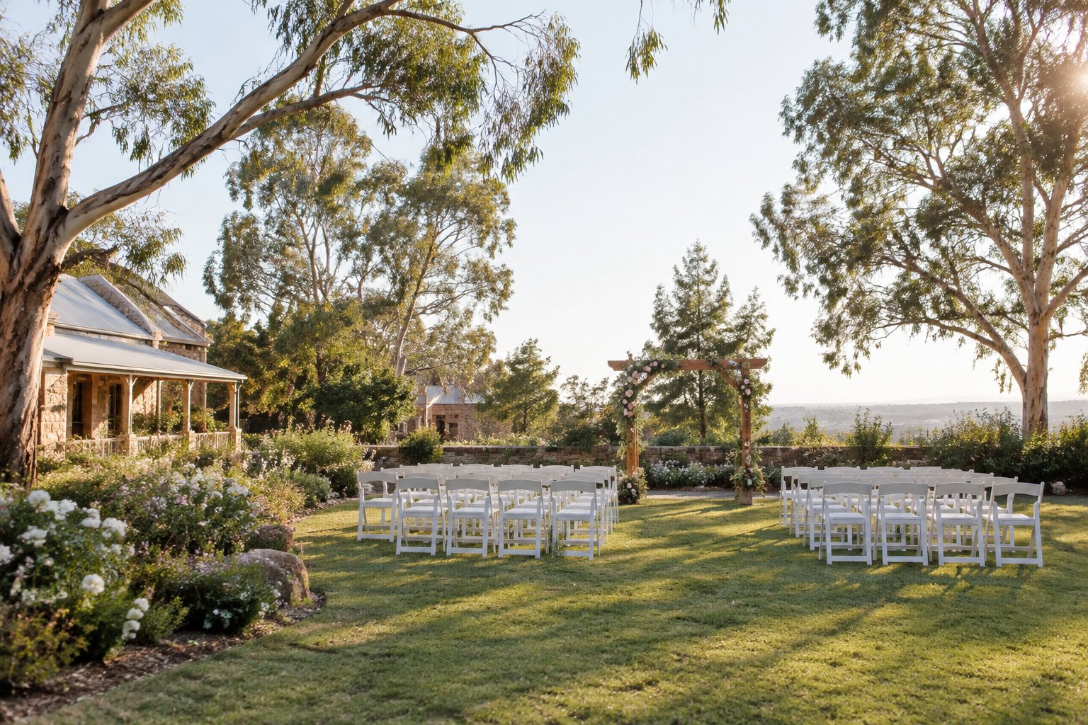 A simple garden wedding venue with white chairs, greenery, and a floral arch in warm sunlight.