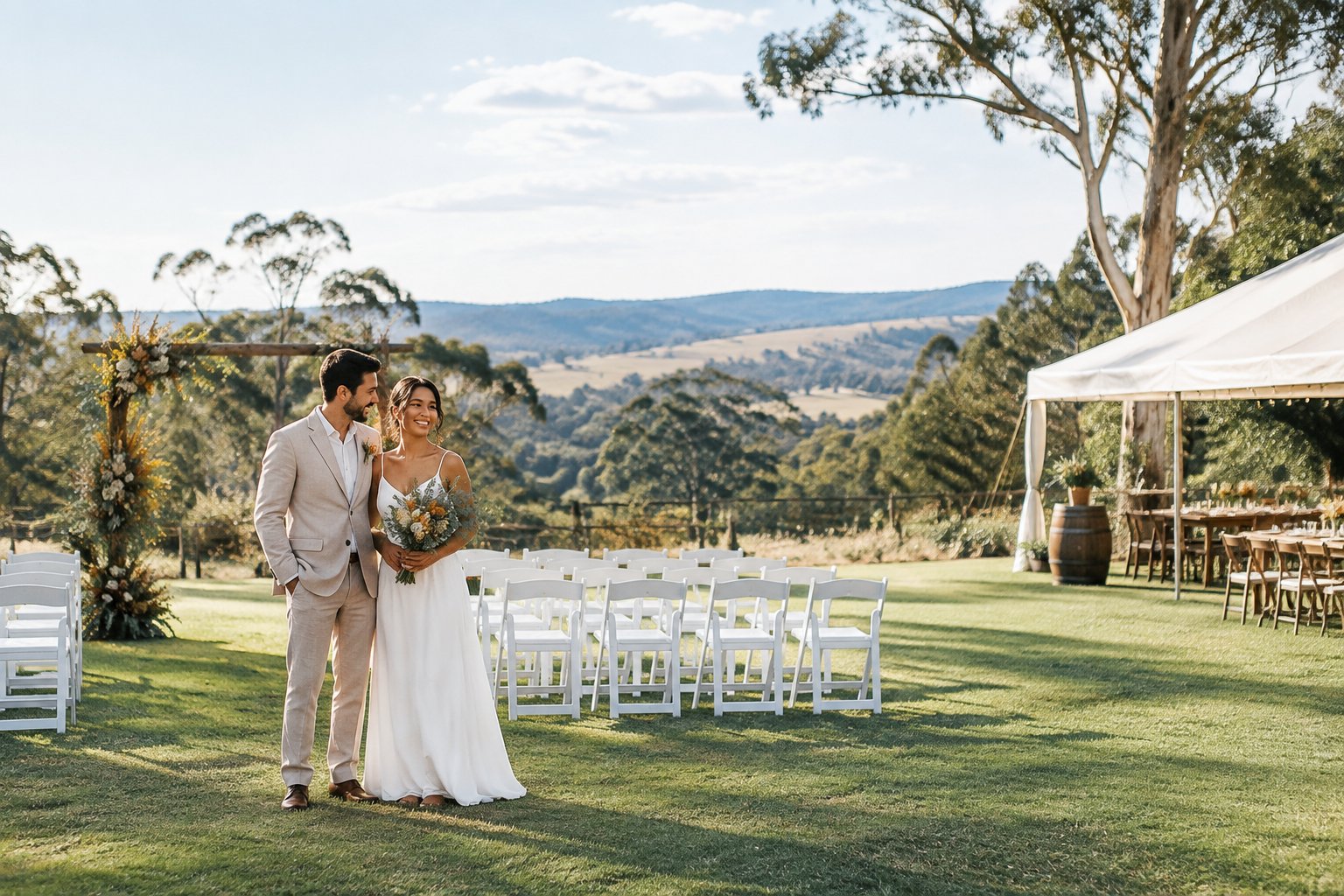 A couple stands beside a simple outdoor wedding ceremony setup in a garden with rolling hills and eucalyptus trees.